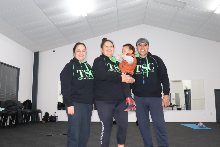 Michelle holds her baby, with partner and daughter beside her. They wear matching sports hoodies and are in an exercise room.