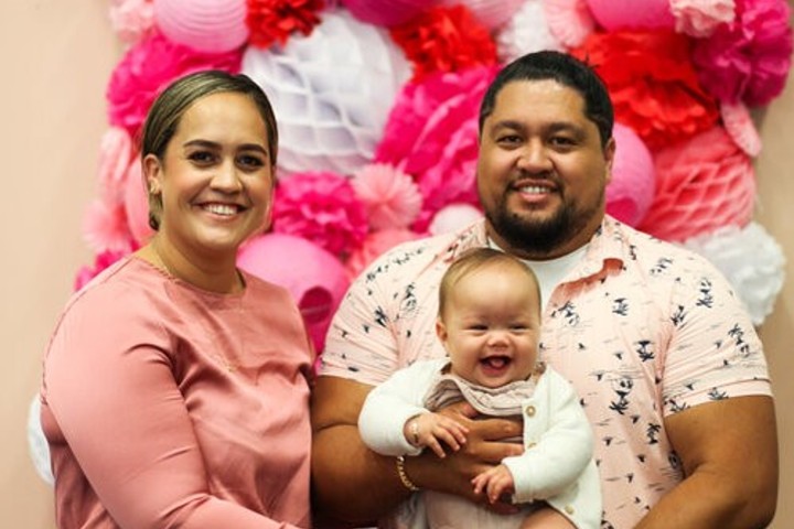 Mo and Zane smiling holding their baby Gray in front of pink, white, and red decorations.